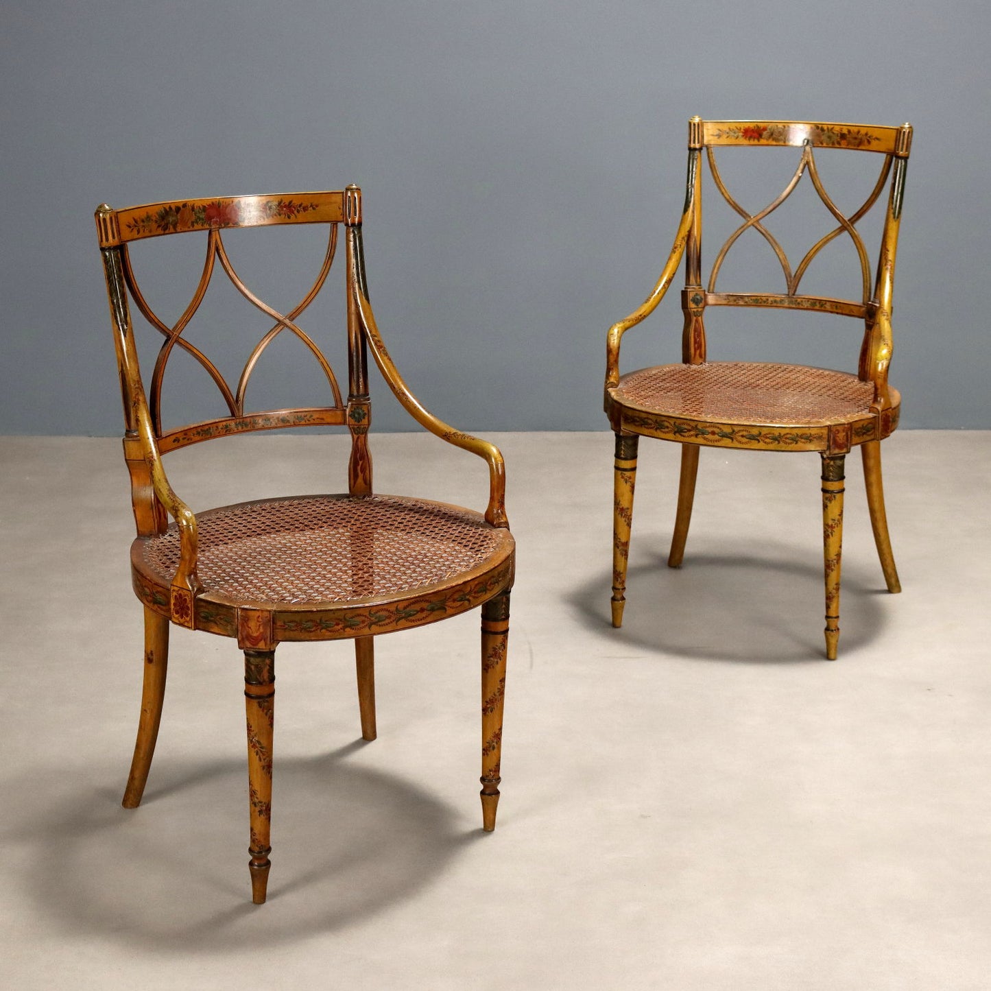 Pair of Regency-style painted armchairs with cane seats, featuring ornate floral hand-painted motifs, X-pattern carved backrests, and slender tapered legs in golden yellow tones.