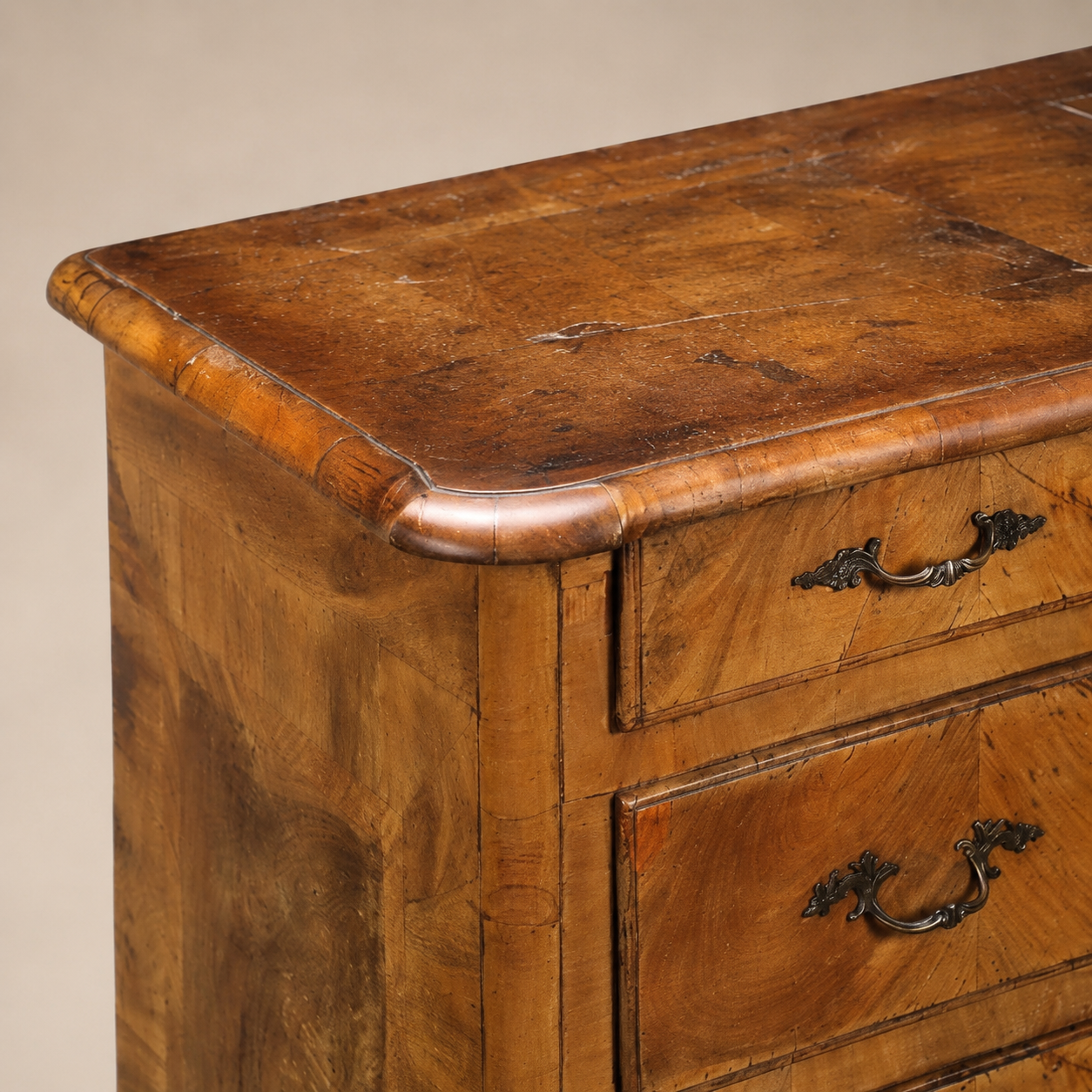 Antique walnut chest of drawers with ornate bronze bail handles, warm honey-brown patina, rounded molded top edge, and rich figured wood grain veneer panels.