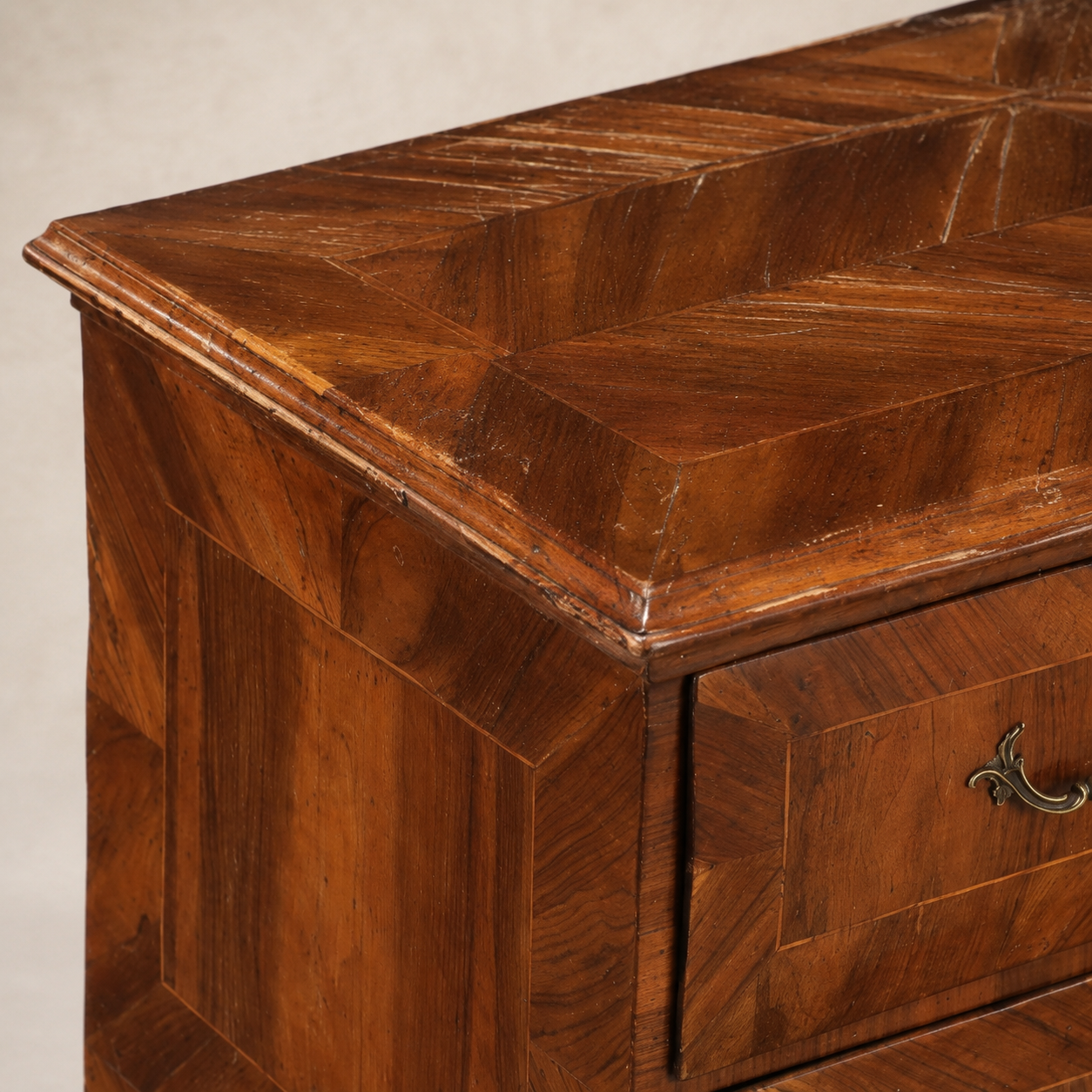 Antique walnut chest of drawers with intricate parquetry top in diamond pattern, curved sides, brass ornate hardware, aged patina showing natural wood grain in warm brown tones.