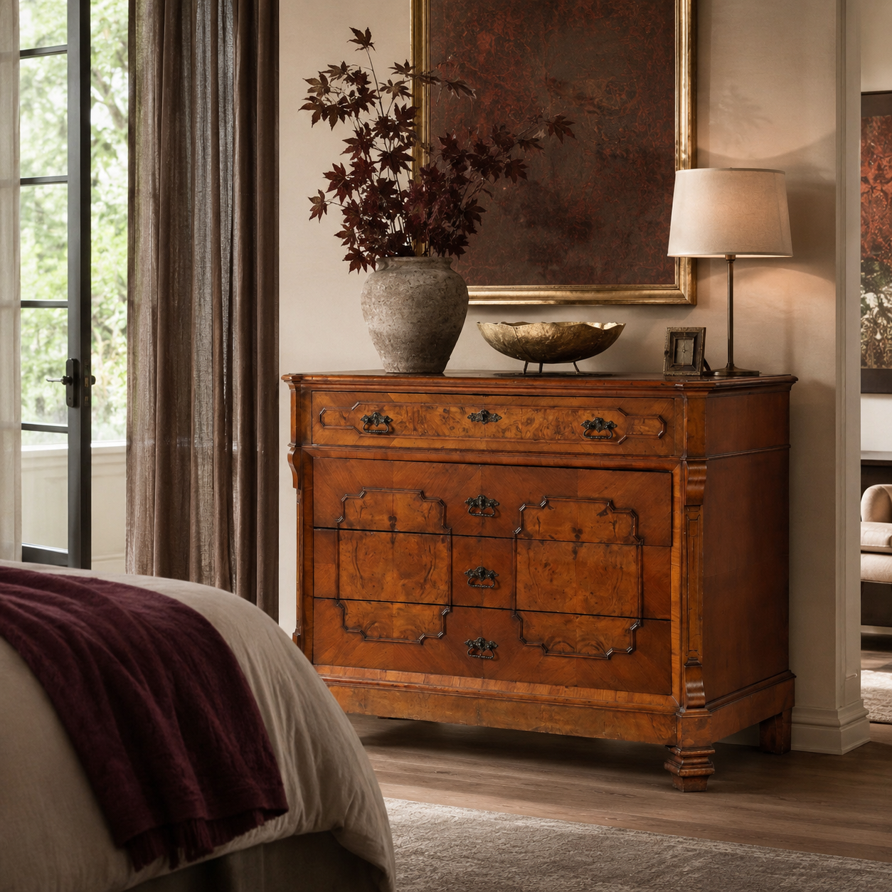 Antique-style burled walnut dresser with ornate brass hardware, carved details, and four drawers in warm honey-brown tones, styled with decorative vase and lamp.