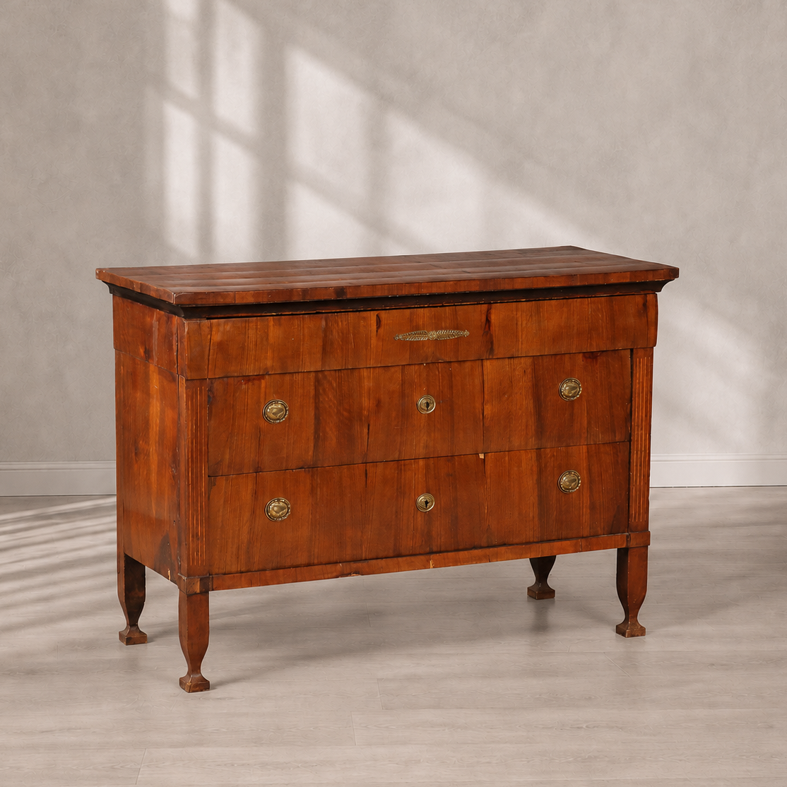 Antique cherry wood chest of drawers with three drawers, brass oval handles and keyhole escutcheons, reeded corner columns, tapered legs, and decorative brass inlay on top drawer.