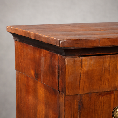 Antique walnut wood chest of drawers with warm honey-brown tones, panel detailing, brass ring pull hardware, and a slightly overhanging flat top with dark molding accent.