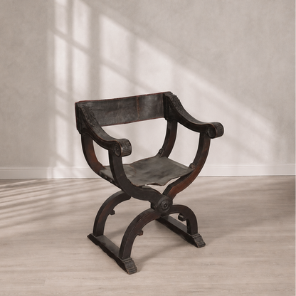 Antique Savonarola X-frame armchair in dark walnut wood with aged black leather seat and back, featuring carved scroll arms and ornate medallion details.