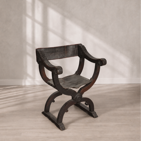 Antique Savonarola X-frame armchair in dark walnut wood with aged black leather seat and back, featuring carved scroll arms and ornate medallion details.