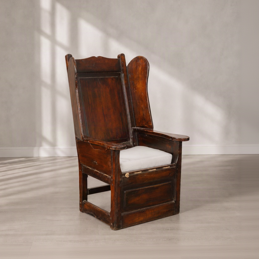 Antique high-back wingback armchair in dark walnut wood with carved panel details, wide flat armrests, stretcher base, and a loose white linen seat cushion.