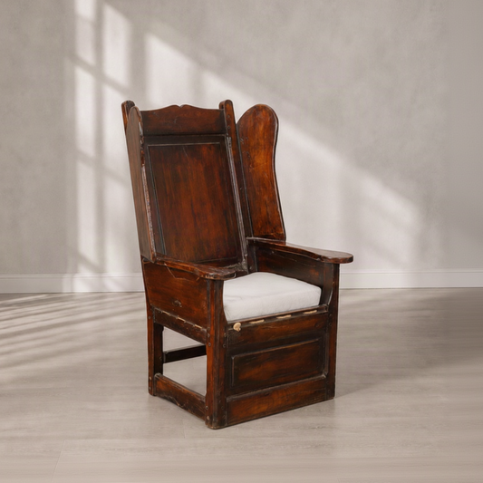 Antique high-back wingback armchair in dark walnut wood with carved panel details, wide flat armrests, stretcher base, and a loose white linen seat cushion.