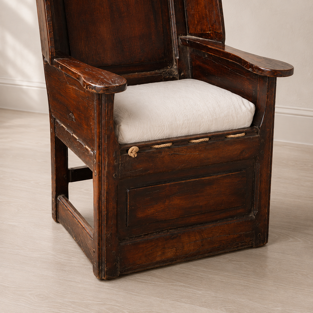 Antique dark brown wooden armchair with high paneled back, worn distressed finish, flat wooden armrests, rope-tied white linen seat cushion, and enclosed panel base storage.