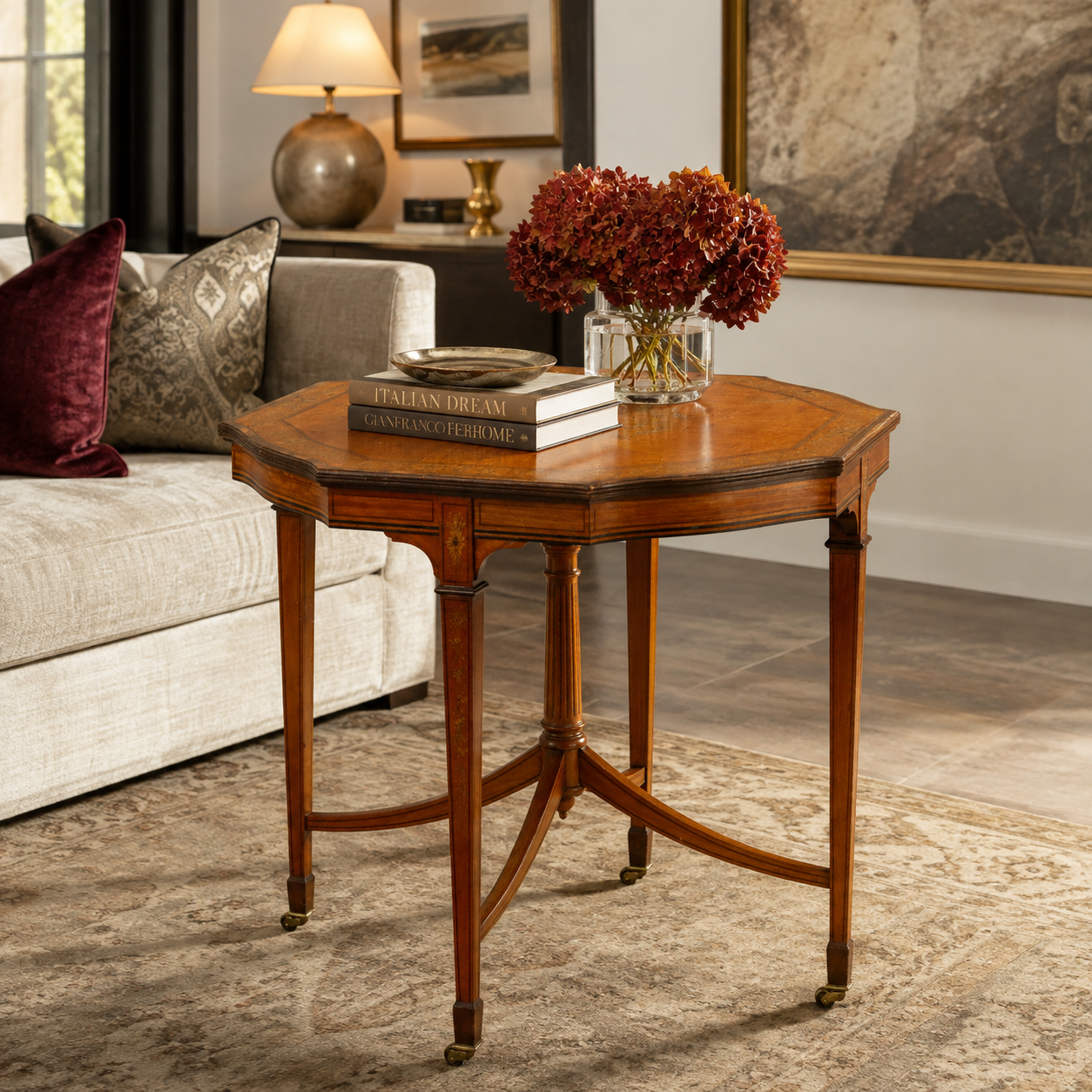 Antique-style octagonal walnut occasional table with inlay details, tapered legs, brass caster wheels, and curved stretcher base, styled with books and red hydrangeas.