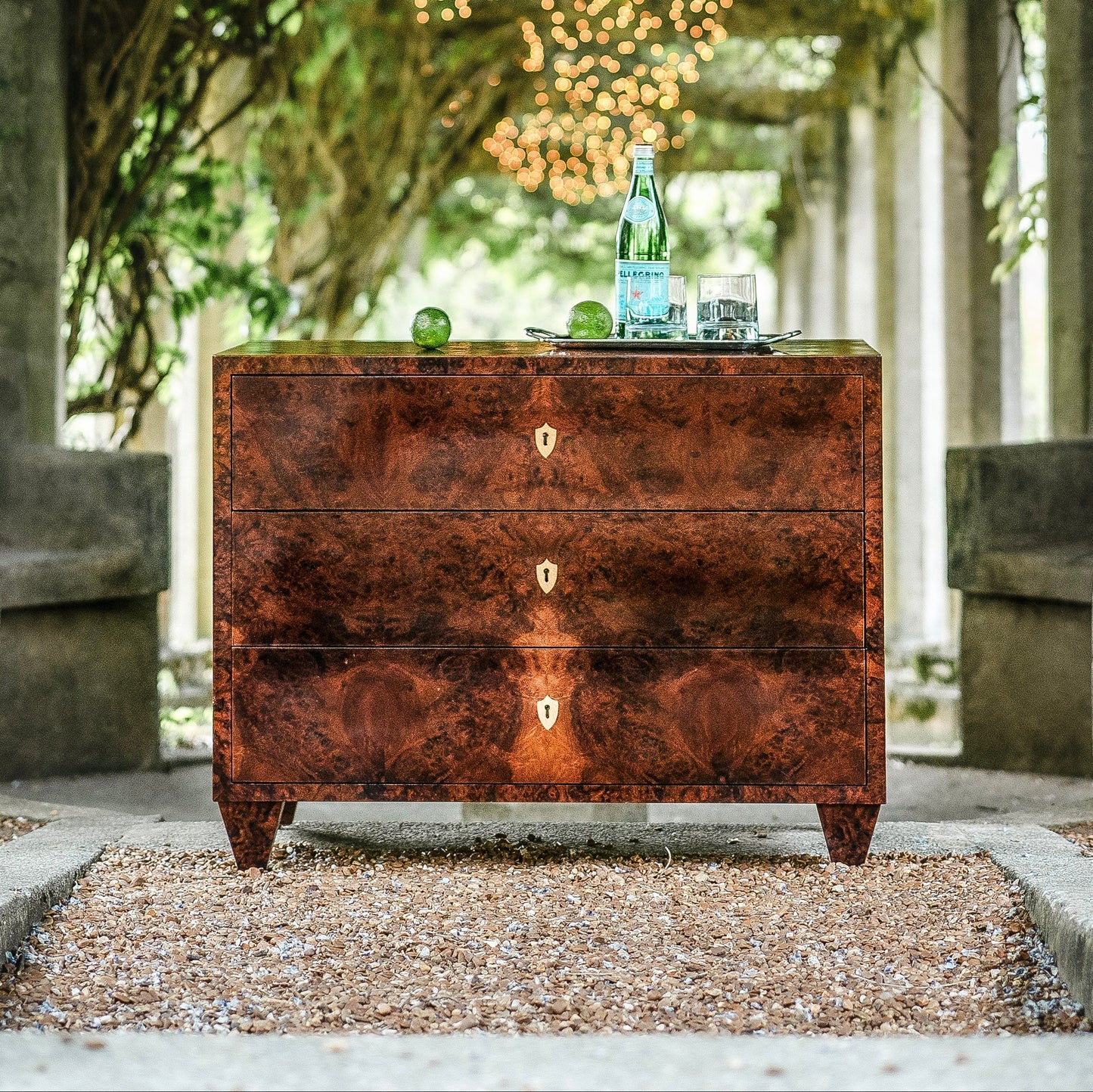 Rich walnut burl veneer chest of drawers with three drawers featuring distinctive wood grain patterns and brass keyhole escutcheons, positioned outdoors on gravel surface