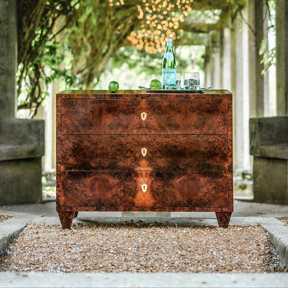 Rich walnut burl veneer chest of drawers with three drawers featuring distinctive wood grain patterns and brass keyhole escutcheons, positioned outdoors on gravel surface