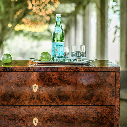 Burl wood chest of drawers with rich brown grain pattern, brass shield-shaped keyholes, two drawer fronts, topped with silver tray holding Pellegrino bottle and glasses