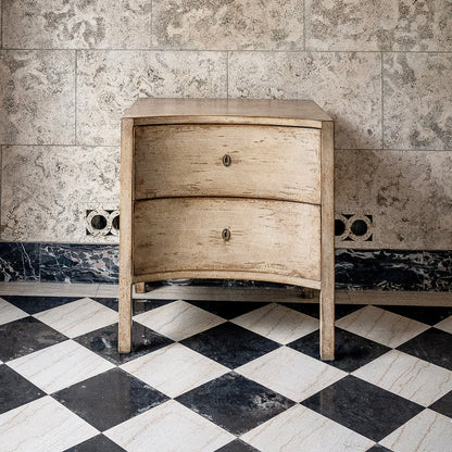Natural wood chest with curved front featuring two drawers and brass oval handles, standing on four legs against marble wall backdrop with black and white checkered floor