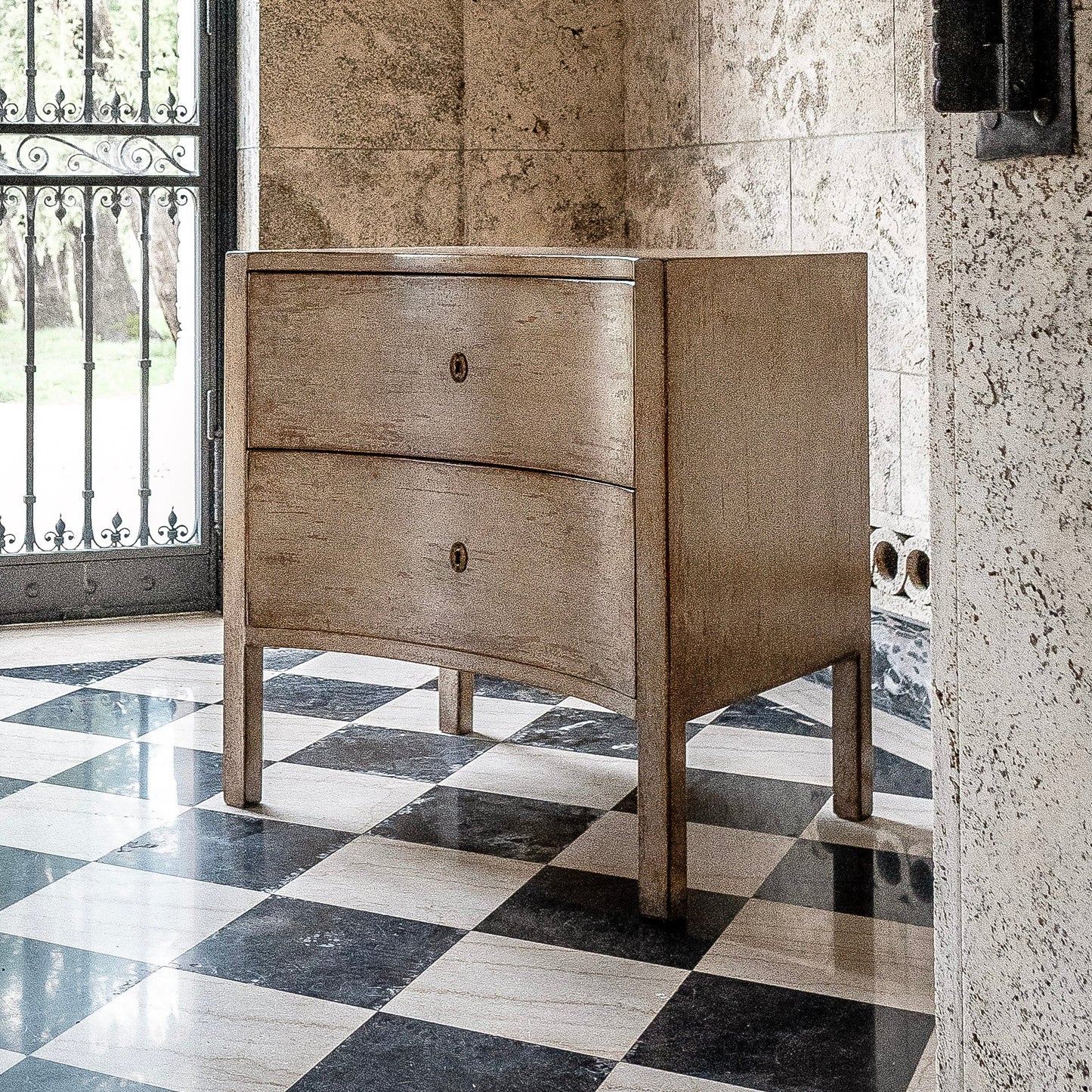 Two-drawer wooden chest with tapered legs and brass hardware, featuring a natural wood finish with visible grain. Positioned on black and white checkered marble floor against stone wall.