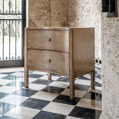 Two-drawer wooden chest with tapered legs and brass hardware, featuring a natural wood finish with visible grain. Positioned on black and white checkered marble floor against stone wall.