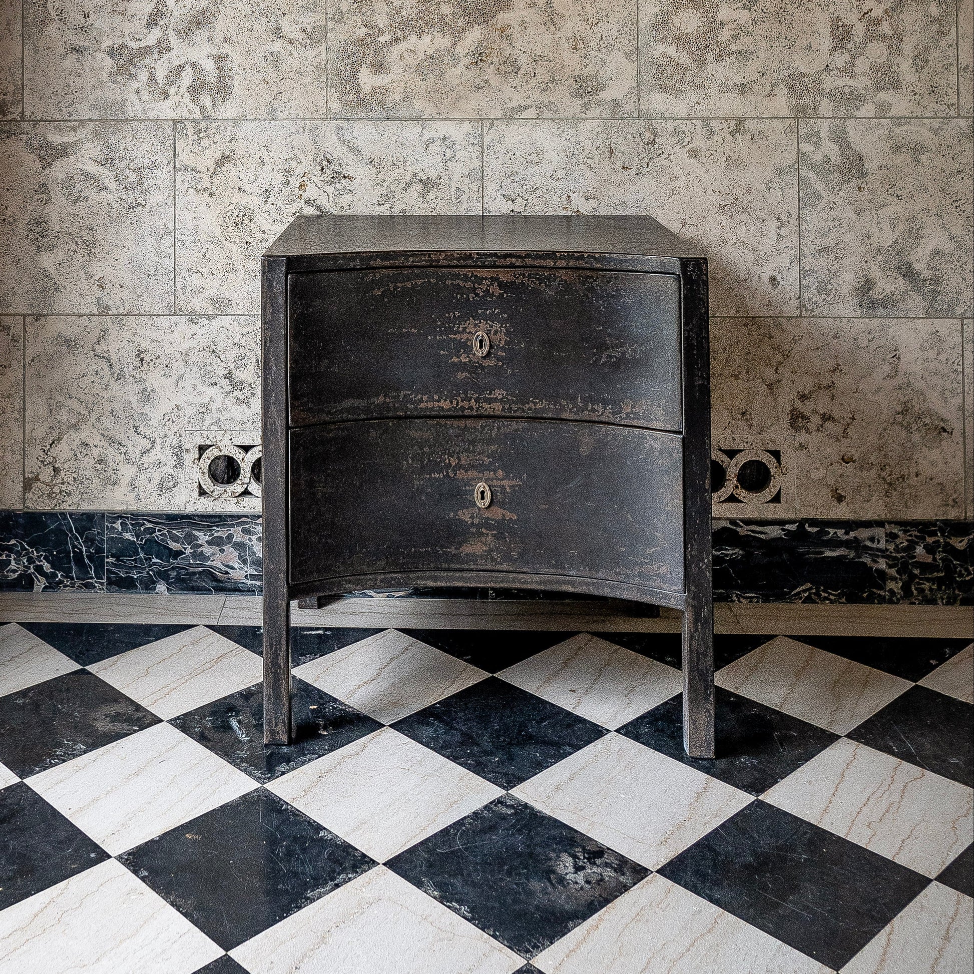 Black distressed wooden chest with two curved drawers and curved legs, featuring weathered finish and brass keyhole hardware, positioned on checkered marble floor