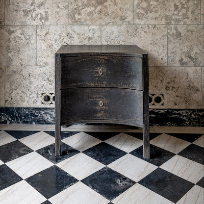 Black distressed wooden chest with two curved drawers and curved legs, featuring weathered finish and brass keyhole hardware, positioned on checkered marble floor