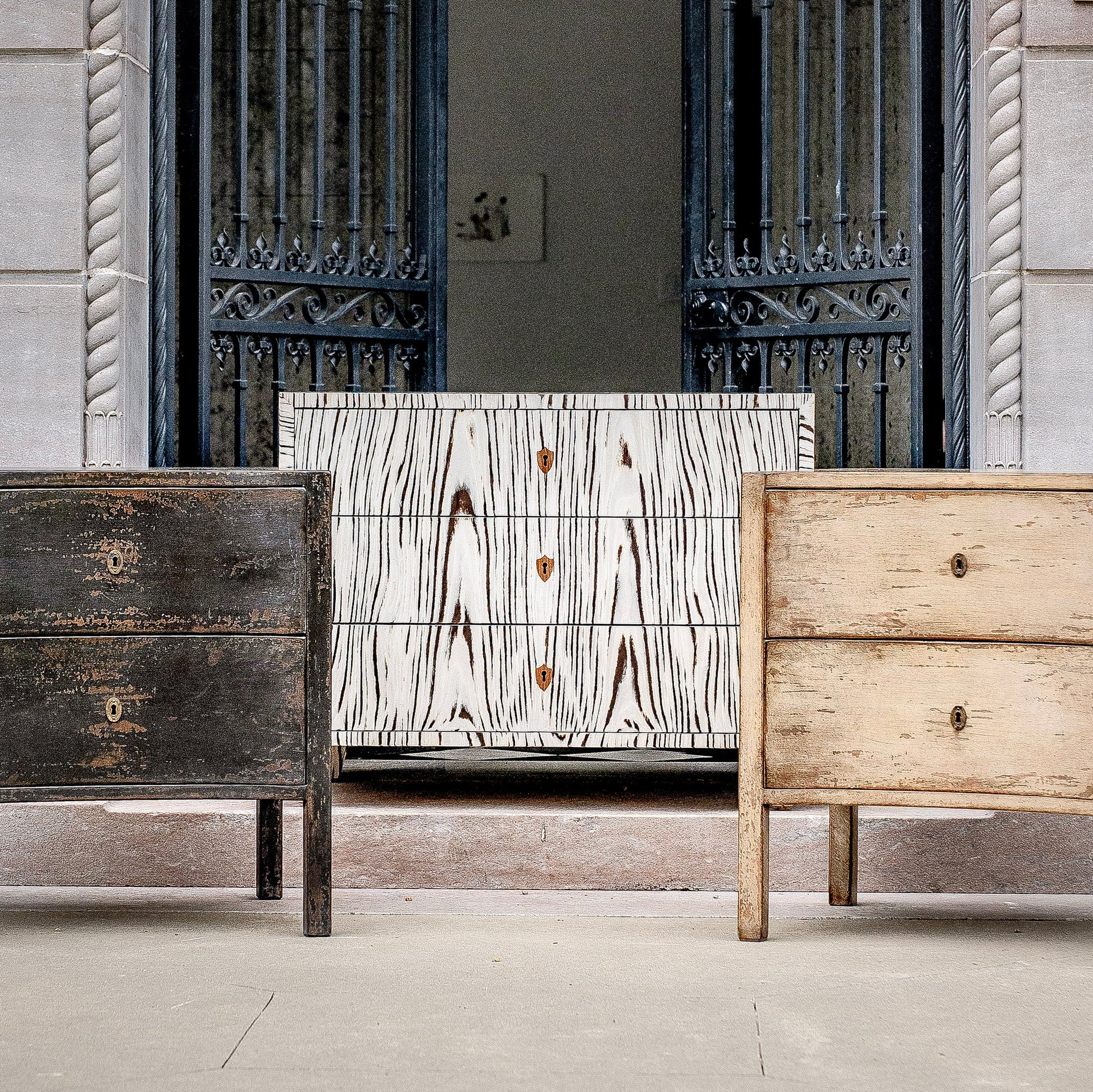 Three chest of drawers displayed together showing different finishes - dark distressed wood on left, white with black wood grain pattern in center, and natural light wood on right