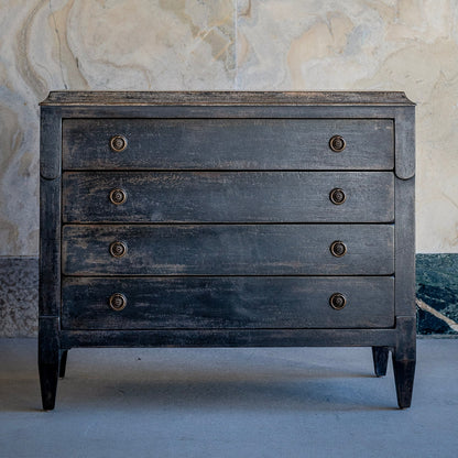 Distressed black wooden chest of drawers with four drawers, bronze circular knob hardware, tapered legs, and weathered finish against marble wall backdrop