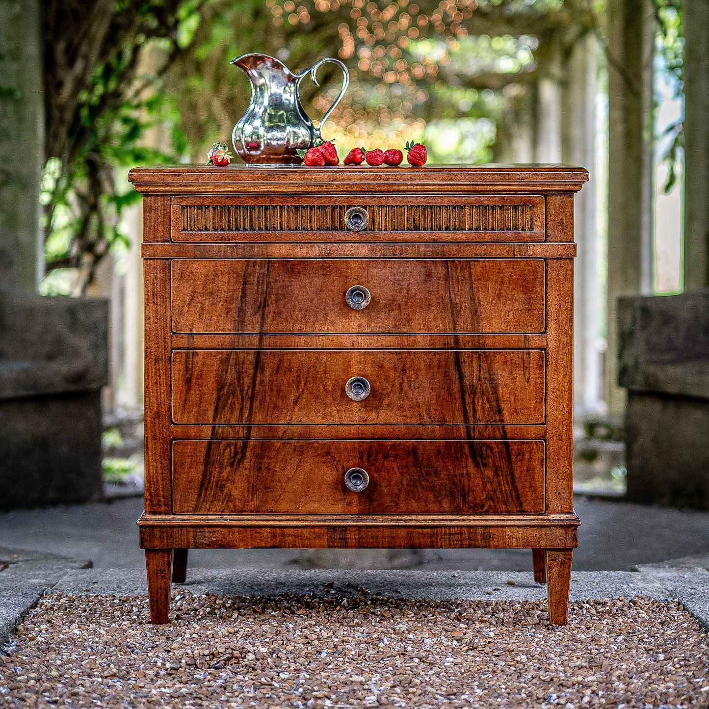 Traditional walnut chest of drawers with four graduated drawers, decorative reeded top drawer, round brass knobs, and tapered legs, styled with silver pitcher and strawberries