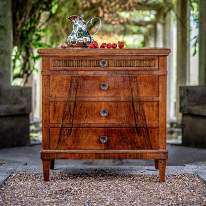 Traditional walnut chest of drawers with four graduated drawers, decorative reeded top drawer, round brass knobs, and tapered legs, styled with silver pitcher and strawberries
