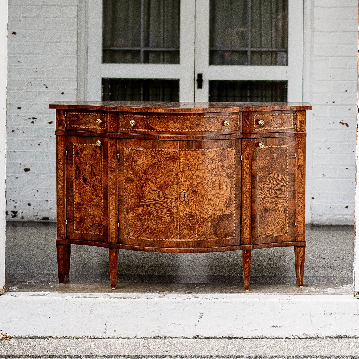 Antique walnut sideboard with curved front, featuring rich burl wood grain patterns, brass hardware, four drawers above cabinet doors, and tapered legs on white porch