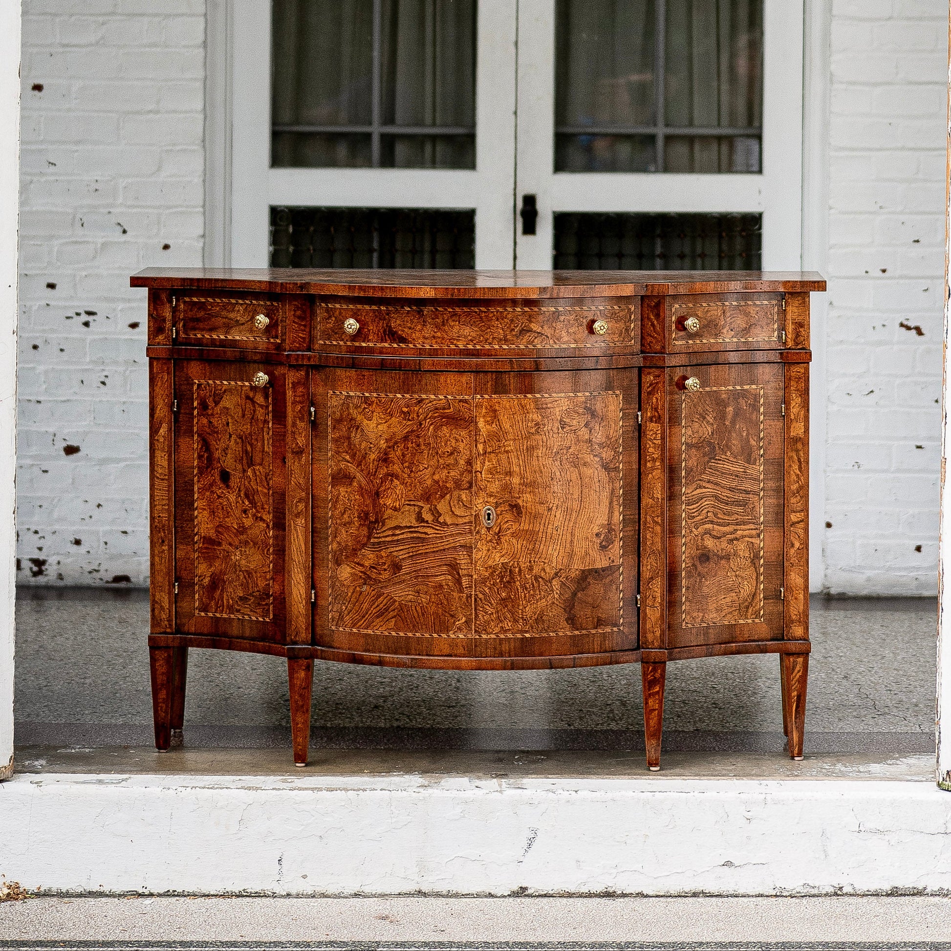 Antique walnut sideboard with curved front, featuring rich burl wood grain patterns, brass hardware, four drawers above cabinet doors, and tapered legs on white porch