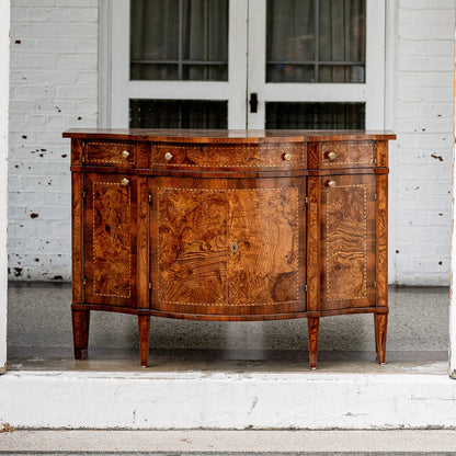 Antique walnut sideboard with curved front, featuring rich burl wood grain patterns, brass hardware, four drawers above cabinet doors, and tapered legs on white porch