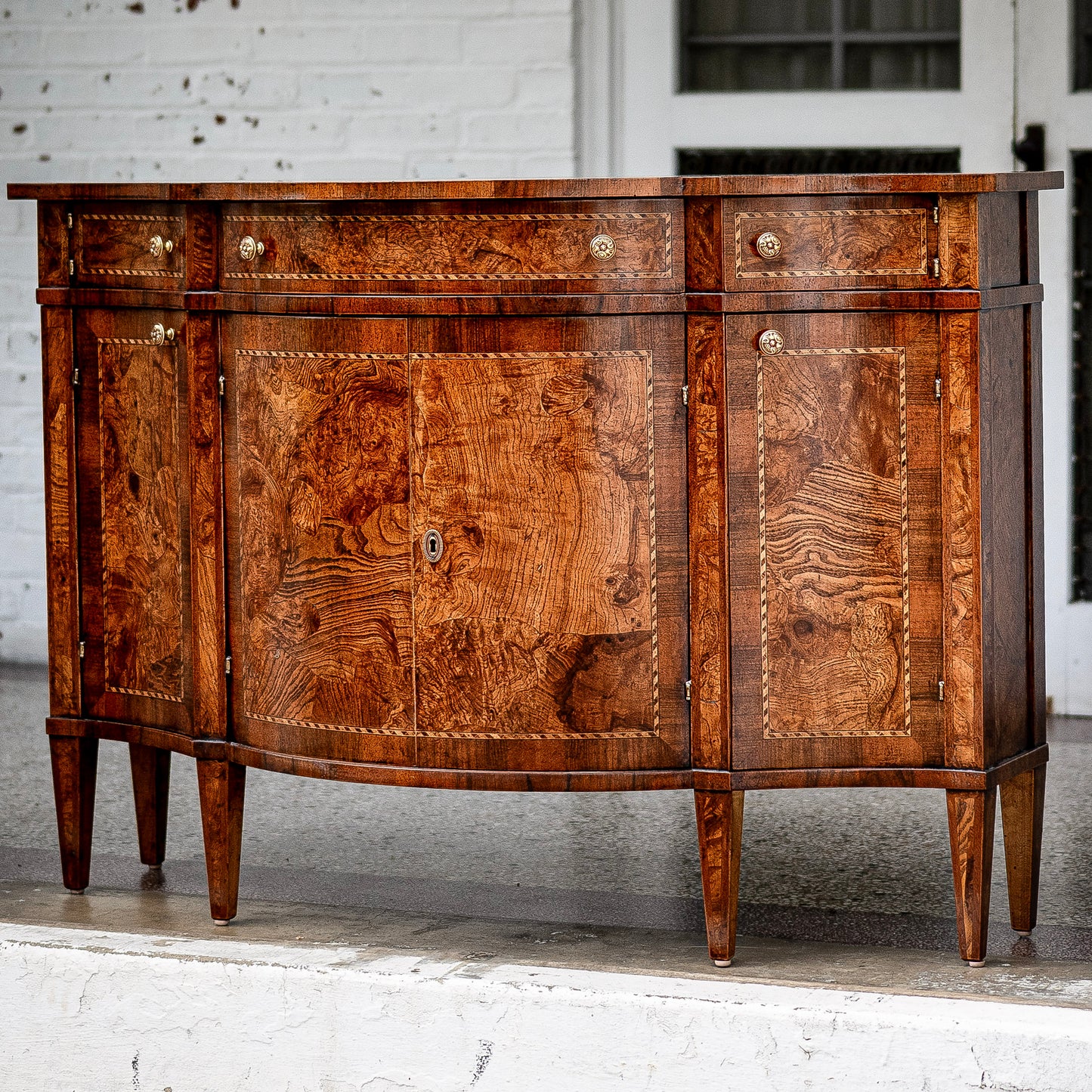Traditional sideboard buffet in rich burled walnut wood with distinctive swirled grain patterns. Features four cabinet doors, three drawers with brass knobs, and tapered legs.