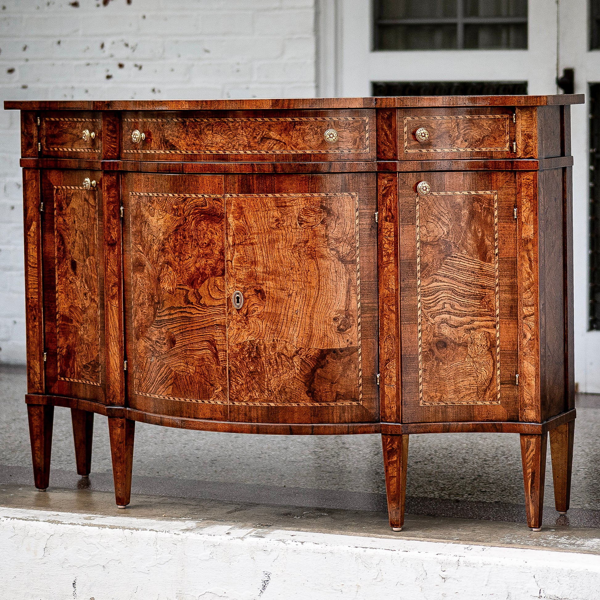 Traditional sideboard buffet in rich burled walnut wood with distinctive swirled grain patterns. Features four cabinet doors, three drawers with brass knobs, and tapered legs.