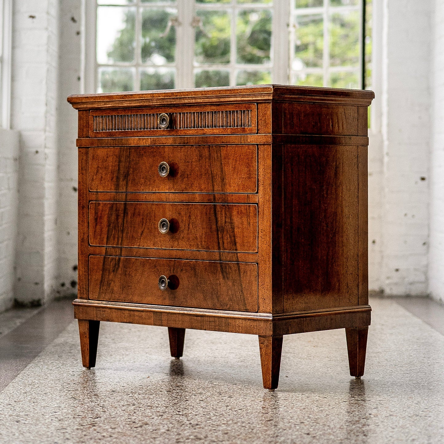 Traditional wooden chest with rich brown finish featuring four drawers including decorative fluted top drawer, brass hardware, and tapered legs on polished concrete floor