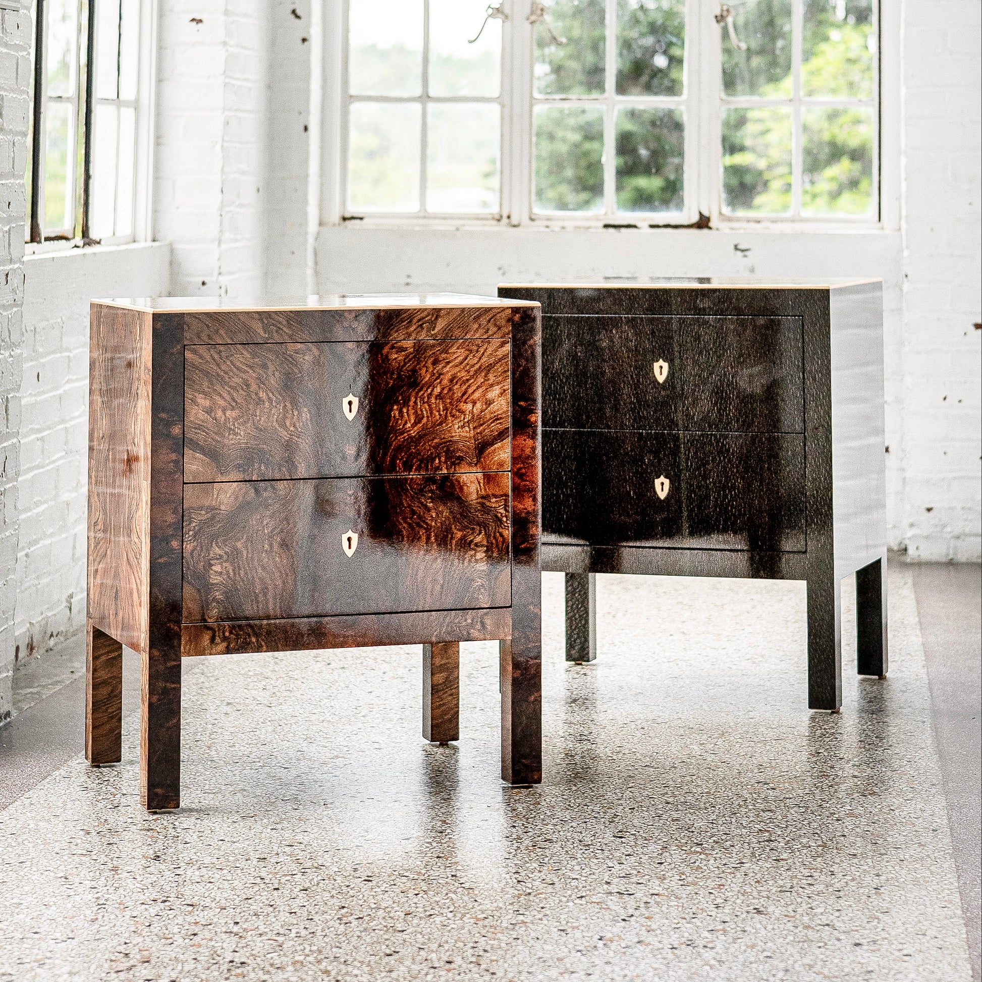 Two wooden nightstand chests with rich wood grain patterns - one in warm brown walnut finish, one in dark ebony black. Both feature two drawers with brass keyhole hardware and sleek legs.