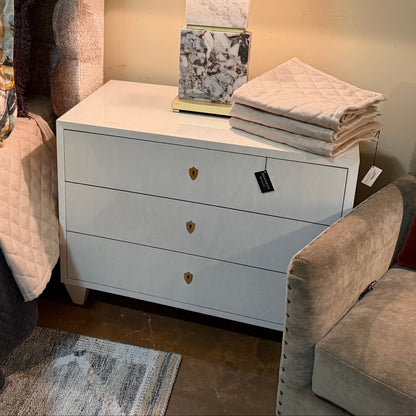 White modern chest of drawers with three drawers featuring brass-colored hexagonal pulls, clean lines, and tapered legs in contemporary furniture showroom setting