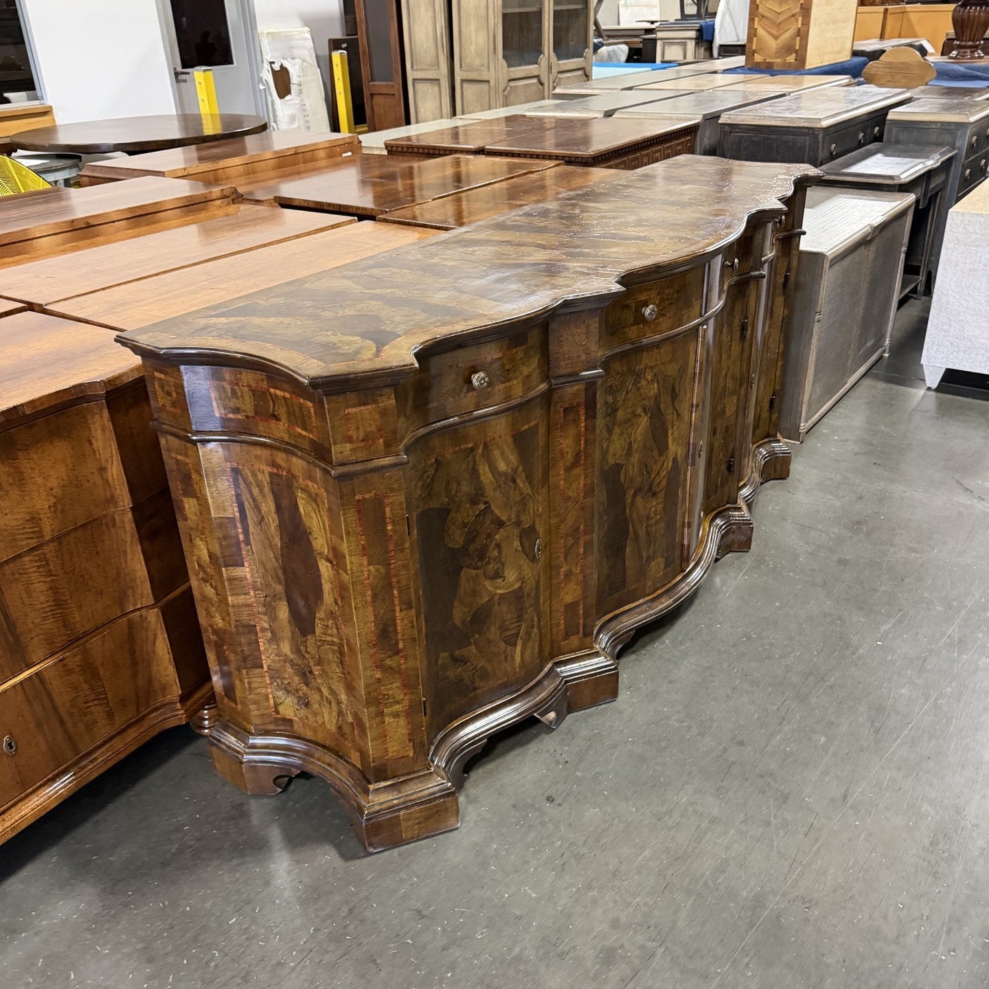 Traditional burl wood sideboard with curved front, featuring rich brown wood grain patterns, ornate molded base, two drawers with round knobs, and decorative curved legs in warehouse setting