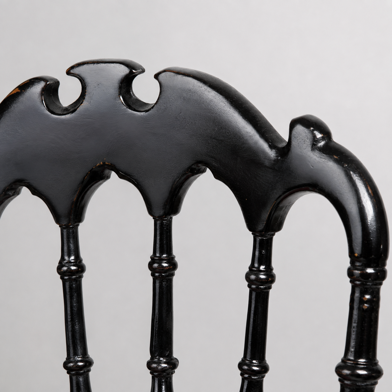 Black ebonized wood chair back detail featuring Gothic arch spindles and decorative carved crest rail with bat-wing silhouette cutouts, distressed finish