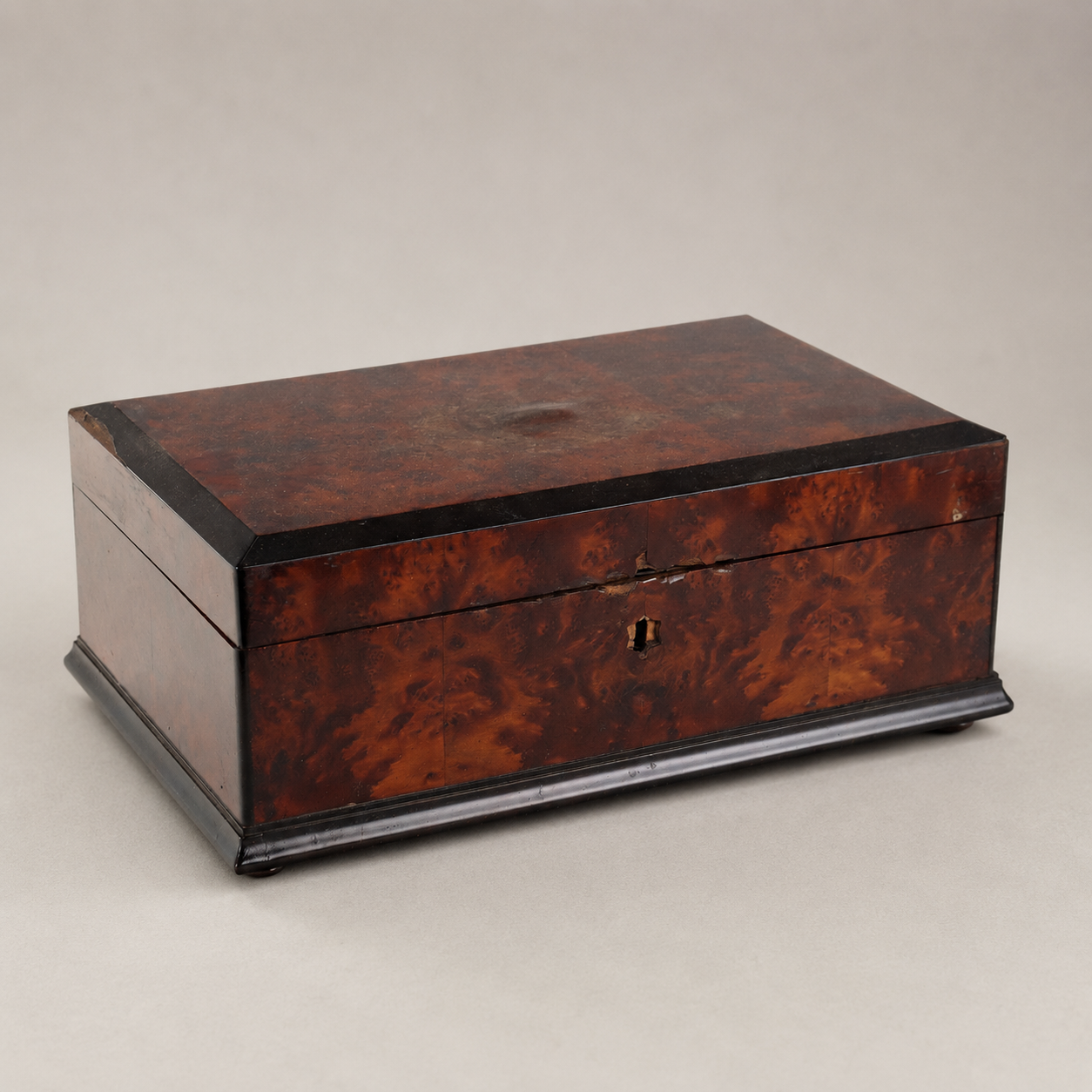Antique burl wood decorative box with ebonized trim, rich reddish-brown swirling grain pattern, keyhole escutcheon on front, raised on small feet with molded base.