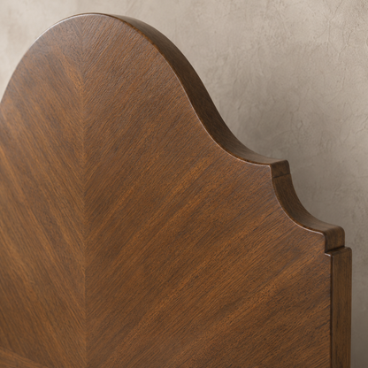 Close-up of a warm walnut-toned wooden bed headboard featuring elegant curved silhouette with bookmatched chevron wood grain veneer and stepped molding detail.