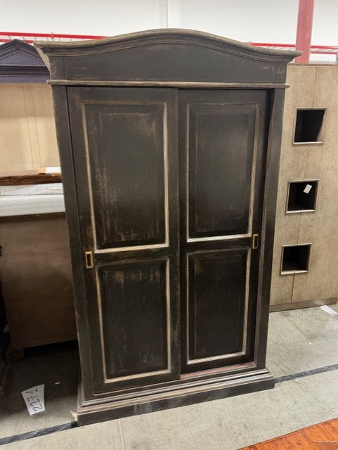 Dark brown traditional armoire with arched top, glass panel doors, and gold-tone hardware. Features classic raised panel design with decorative molding trim in showroom setting.
