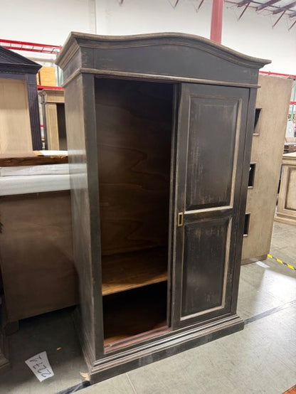 Dark gray traditional armoire with curved crown molding, featuring one open door revealing interior shelving and one closed door with raised panel design, displayed in warehouse setting
