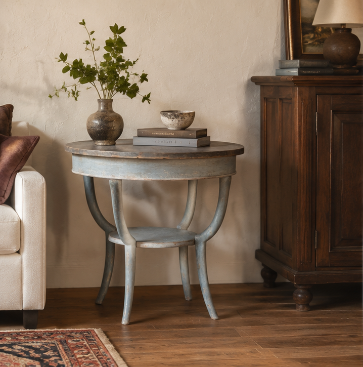 Round occasional table with curved legs in gray-blue finish, featuring distressed paint treatment. Styled with vase of greenery and books on wooden floors beside upholstered chair.