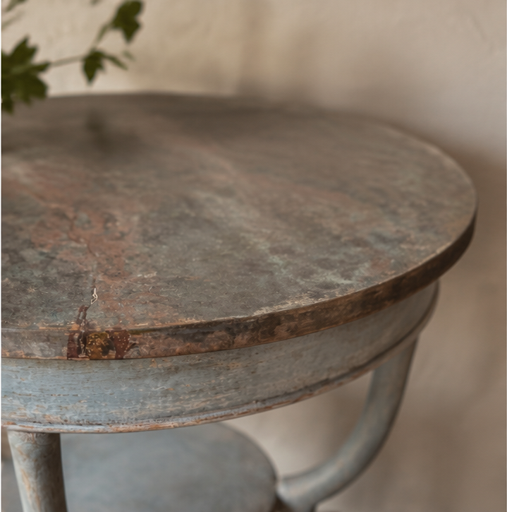 Round weathered occasional table with distressed gray-brown painted finish showing natural wood undertones. Features curved pedestal base with vintage farmhouse styling and rustic patina.