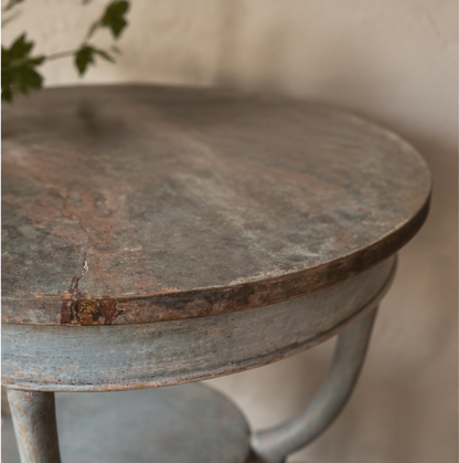 Round weathered occasional table with distressed gray-brown painted finish showing natural wood undertones. Features curved pedestal base with vintage farmhouse styling and rustic patina.